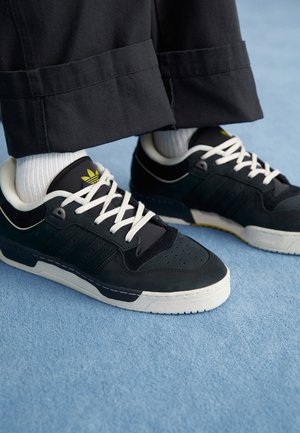 Sorte og mørkegrønne sneakers med hvide snørebånd, tekstureret ruskind og læderdetaljer, lavt design, med et gult logo og hvid sål.