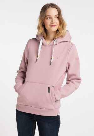Jonge vrouw draagt een lichtroze hoodie met witte trekkoorden en donkere spijkerbroek, handen in de voorzak, kijkt iets naar de zijkant en glimlacht.
