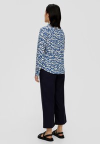 Langärmliges Hemd mit einem blauen und weißen abstrakten Muster, kombiniert mit dunkelmarineblauen, knöchellangen Hosen und schwarzen, geflochtenen Sandalen.