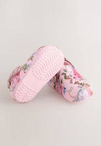 Zoccoli rosa da bambino senza lacci con motivi floreali e farfalle, uno che mostra la suola texturizzata, su uno sfondo neutro.