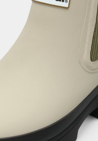 Μπεζ λαστιχένια μπότα με λεία επιφάνεια, που διαθέτει υφασμάτινες πράσινες ελαστικές πλευρικές επενδύσεις και μαύρη σόλα. Το λογότυπο είναι ορατό στο πάνω μέρος.