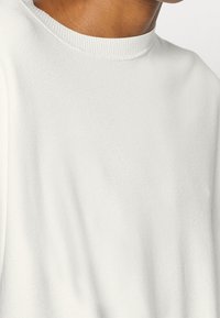 Bílý pletený svetr s žebrovaným kulatým výstřihem a hladkou texturou, který odhaluje část límce a ramenní oblasti.