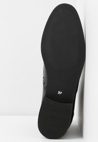 Semelle de chaussure noire avec des motifs de grip texturés, taille numéro 37 imprimée en blanc près du talon, positionnée debout contre une surface blanche.