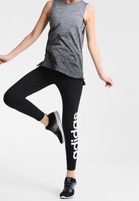 Mustat urheiluleggingssit, joissa on valkoinen "adidas"-logo, yhdistettynä harmaaseen hihattomaan paitaan. Mallilla on päällään mustat ja harmaat urheilukengät.