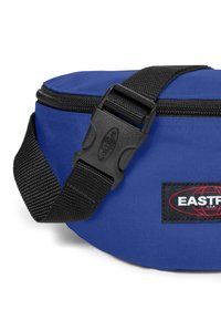 Modrý látkový pasový vak se zipovým uzávěrem, s černým nastavitelným popruhem a plastovou přezkou. Obsahuje označení značky na boku.