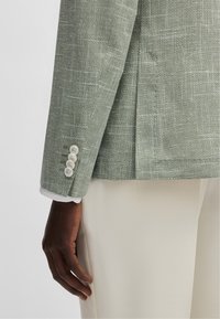 Blazer texturé vert clair avec des accents blancs, fente simple, et quatre boutons blancs sur le poignet. Pantalons de couleur crème visibles en dessous.