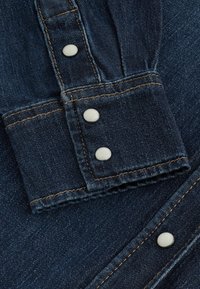 Tessuto di denim blu scuro con un orlo piegato che presenta due bottoni a pressione metallici e cuciture a contrasto. La trama è liscia con una tessitura visibile.