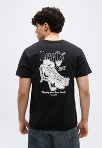 Νέος άνδρας που φοράει μαύρο μπλουζάκι Levi's με καρτούν χοτ ντογκ, καθισμένος αναπαυτικά σε καρέκλα, με το κείμενο «Κρατώντας τα ψωμάκια σου ζεστά από το 1873» στην πλάτη.