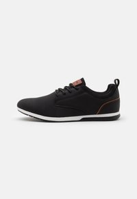 Zwarte sportieve schoenen met een gladde synthetische bovenkant, witte tussenzool en een bruin leren accent op de hiel; voorzien van vetersluiting en een hulpmiddel voor gemakkelijk aantrekken.