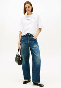 Camiseta blanca extragrande con logo impreso, jeans azules de talle alto, bolso negro y zapatos planos negros brillantes, posando frente a un fondo liso.