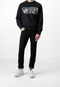 Svart sweatshirt med tryckt logotyp "Versace Jeans Couture", tillsammans med svarta jeans och vita sneakers. Mjukt tyg, avslappnad passform.