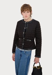 Svart quiltet jakke med rund hals, trykknapper og lærdetaljer, kombinert med lyseblå jeans og en svart håndveske.