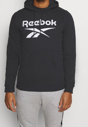 Man draagt een zwarte Reebok-hoodie met wit logo en grijze joggingbroek, staand tegen een effen witte achtergrond.