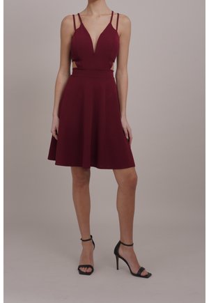 Femme portant une robe bordeaux sans manches avec un décolleté en V profond, des côtés ajourés, et des escarpins noirs à brides à la cheville, debout devant un fond uni.