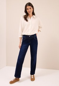 Mujer de pie con la mano en el bolsillo, vistiendo una camisa blanca de botones, jeans azul oscuro y zapatillas marrones, frente a un fondo beige.