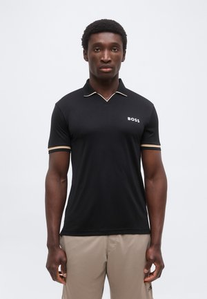 Homme portant un polo noir BOSS à manches courtes avec une bordure beige et un pantalon beige, debout devant un fond clair uni.