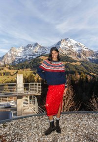 Jupe rouge en sequins avec fente latérale, pull en maille bleu marine avec motif multicolore, et bottines marron. Décor de montagne en plein air.