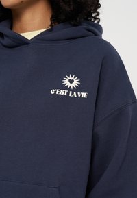 Personne portant un sweat à capuche bleu marine avec un graphisme blanc de soleil et de cœur au-dessus du texte "C'EST LA VIE" sur la poitrine gauche.