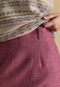 Różowe spodnie w jodełkę o gładkiej fakturze i dyskretnej kieszeni. Wzorzysty sweter z dzianiny częściowo zasłania pas spodni.