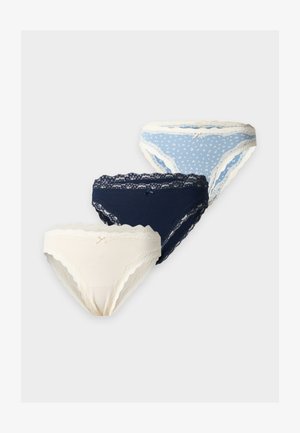 Donna in piedi scalza che indossa un reggiseno bianco e mutandine blu a pois con bordo di pizzo, su uno sfondo chiaro uniforme.