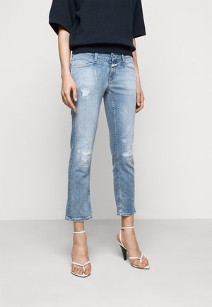 Henkilö, jolla on vaaleansiniset kulutetut lyhyet farkut ja valkoiset korkokengät, seisoo yksinkertaisen taustan edessä.