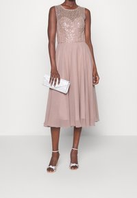 En utsvängd klänning till knäna med en paljettbeströdd beige överdel och en mjuk, flödande kjol i en dov rosa nyans. Matchas med silverfärgade remsandaler och en liten handväska.