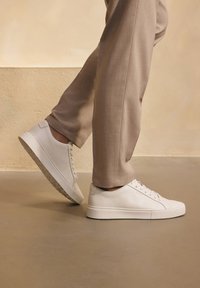 Witte leren sneakers met gestreepte zolen, ronde vorm en platte veters. Gecombineerd met beige op maat gemaakte broeken, staand op een gladde ondergrond.
