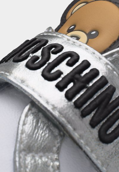 Ασημένιο λουράκι παπουτσιού με ανάγλυφο μαύρο κείμενο "MOSCHINO" πάνω σε γραφικό καφέ αρκούδας στην εσωτερική σόλα του παπουτσιού.