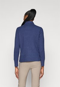 Chaqueta ajustada de color azul marino con cuello alto y mangas largas. Presenta detalles de costura y una tela texturizada. Combinada con pantalones en tonos neutros.