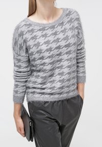 Donna che indossa un maglione grigio in tessuto a scacchi e pantaloni di pelle nera, mentre tiene una pochette di pelle nera nella mano sinistra.