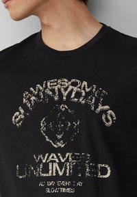 Zwarte katoenen t-shirt met een versleten crèmekleurige graphic en de tekst "AWESOME SLEAZY DAYS" en "WAVES UNLIMITED" op de voorkant.