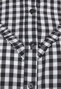 Camicia a scacchi bianca e nera con chiusura a bottoni, dettagli arricciati sulle spalle e un motivo a scacchi su tutta la superficie.