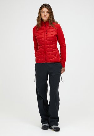 Jonge vrouw staand, gekleed in een rode doorgestikte jas, zwarte outdoorbroek met aantrekkoorden en grijze wandelschoenen tegen een witte achtergrond.