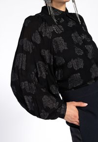 Black blouse with metallic silver abstract pattern, long puffed sleeves, button collar, and sheer fabric, paired with a dark waistband.