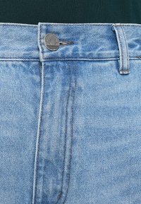 Primo piano della cintura di jeans in denim azzurro chiaro con un bottone metallici che presenta un logo a trifoglio, indossati con una maglia verde scuro.