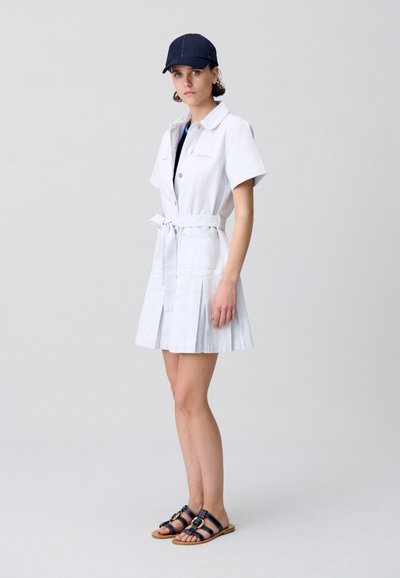 Robe blanche à boutons avec des manches courtes et une ceinture, dotée de deux poches avant, associée à une casquette bleue et des sandales noires avec des accents dorés.