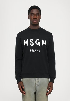 Black long-sleeve sweatshirt with “MSGM MILANO” printed in white worn by person standing against plain white background.