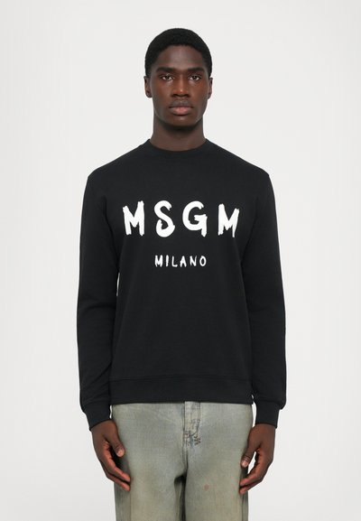 Schwarzes Langarm-Sweatshirt mit der weißen Aufschrift "MSGM MILANO", getragen von einer Person, die vor einem schlichten weißen Hintergrund steht.