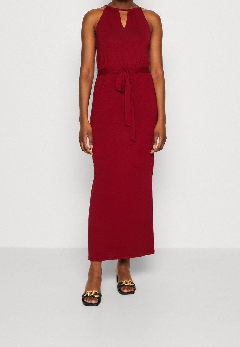 Robe maxi bordeaux à col halter avec une découpe en trou de clé, taille cintrée et ceinture en tissu assortie. Portée avec des sandales noires ornées d'accents dorés.