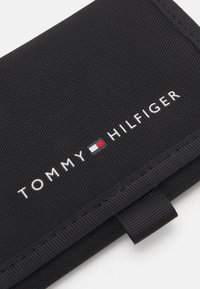 Portefeuille en tissu noir avec le logo "TOMMY HILFIGER" en texte blanc. Surface texturée avec un bord lisse et une petite languette.
