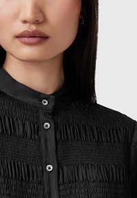 Čierna blúzka s nariasaním, ktorá má golierik, malé strieborné gombíky a textil s vertikálnymi skladmi.