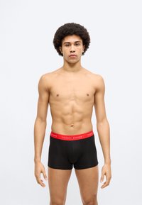 Μαύρα ανδρικά εσώρουχα μπόξερ με κόκκινη ζώνη μέσης που φέρει το κείμενο «TOMMY HILFIGER». Το μοντέλο έχει καλογυμνασμένο σώμα και αφροαμερικανικό χτένισμα.