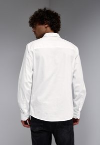Camisa de algodón blanca con mangas largas, puños abotonados y un dobladillo curvado, con un canesú en la parte posterior y una textura suave.