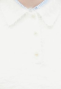 Biely chlpatý sveter s vlnitým golierom a tromi malými gombíkmi, nosený cez golier modro-bielo pruhovanej košele.