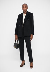 Μαύρο διπλό πετάκι με φαρδιές πέτες, σε συνδυασμό με κρέμα πουκάμισο και μαύρο προσαρμοσμένο παντελόνι. Το μοντέλο κρατάει μια μικρή μαύρη τσάντα.