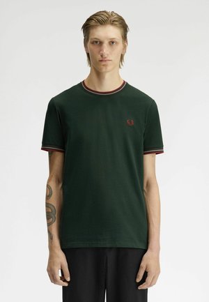 Persona joven con cabello claro, vistiendo una camiseta verde oscuro con ribetes rojos y un pequeño logo rojo en el pecho, de pie frente a un fondo liso.