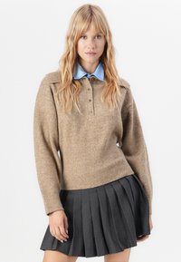Brun strikkegenser med ribbestrikket kant og tre knapper, sammen med en lyseblå skjorte med krage og en mørk plissert skjørt.