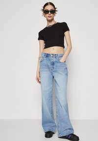 Camiseta negra cortada, jeans de mezclilla de pierna ancha de color azul claro y zapatos negros. El conjunto presenta un collar de cadena plateada y gafas de sol.