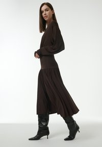 Langärmliges, braunes Kleid mit tailliertem Oberteil und fließendem Rock. Das Model trägt hohe schwarze, spitze Stiefel. Glatter Stoff mit drapierter Textur.