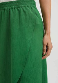 Grüne, texturierte Rock mit einem elastischen Bund, der aus weichem, zerknittertem Stoff besteht und ein Wickeldesign aufweist, das einen sanften Fall und ein angenehmes Finish präsentiert.